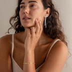 A woman with long curly hair wears Pearl Bow Earrings, a gold bracelet, and a ring as she gazes thoughtfully—showcasing elegant jewelry and timeless style.