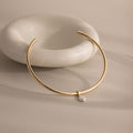 The Pearl Cuff Choker Necklace features a gold band with a delicate single pearl pendant, elegantly presented in front of a white ceramic dish.