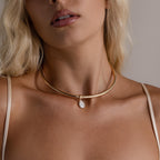A woman with light hair and neutral-toned clothing wears the Pearl Cuff Choker Necklace, featuring a gold band with a single baroque pearl pendant.