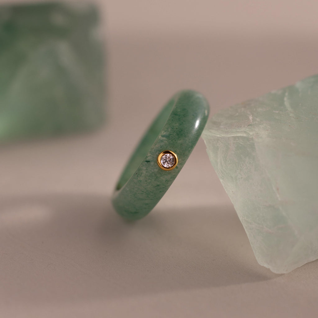 The Jade Diamond Ring showcases a green jade stone and a small diamond set in a gold bezel, presented between two green crystals.