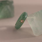 The Jade Diamond Ring showcases a green jade stone and a small diamond set in a gold bezel, presented between two green crystals.