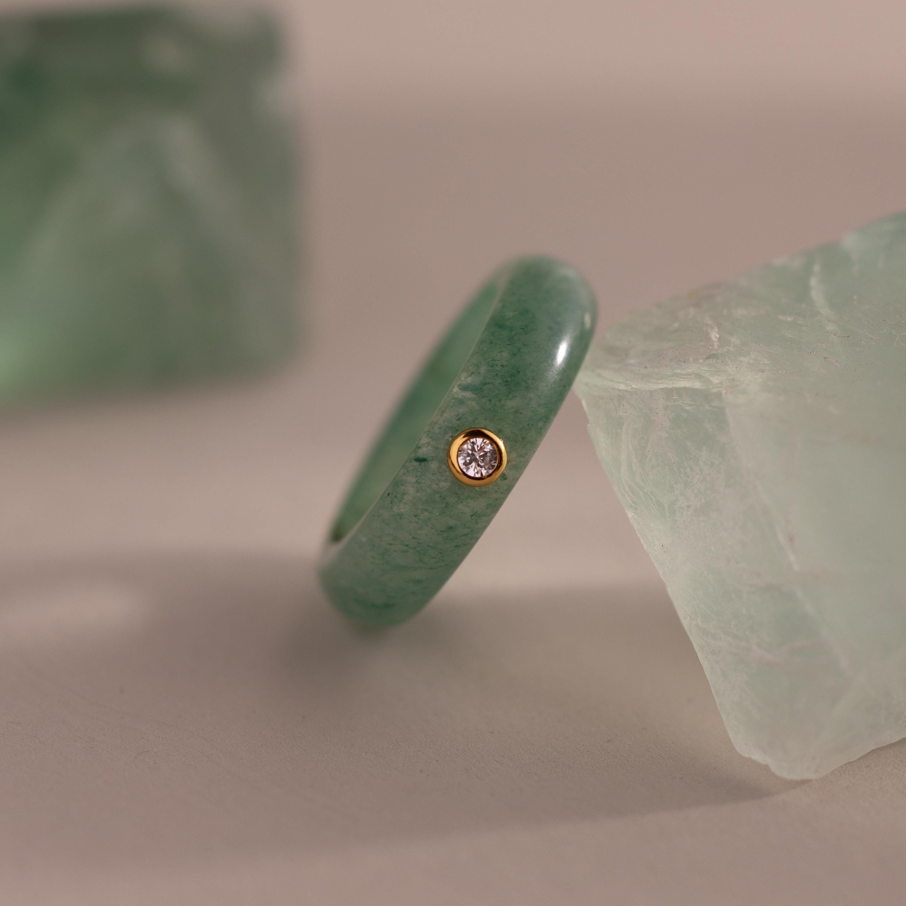 The Jade Diamond Ring showcases a green jade stone and a small diamond set in a gold bezel, presented between two green crystals.