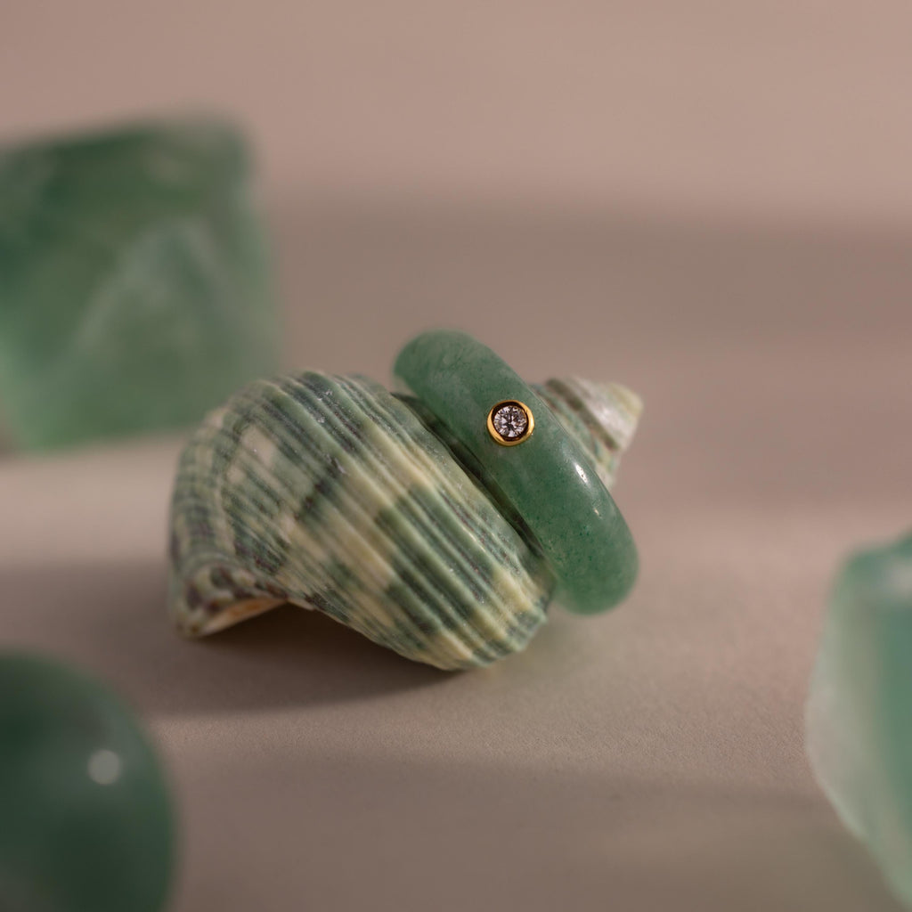 The Jade Diamond Ring features a green jade gemstone and a small diamond set in gold, displayed on a striped seashell with surrounding green stones.