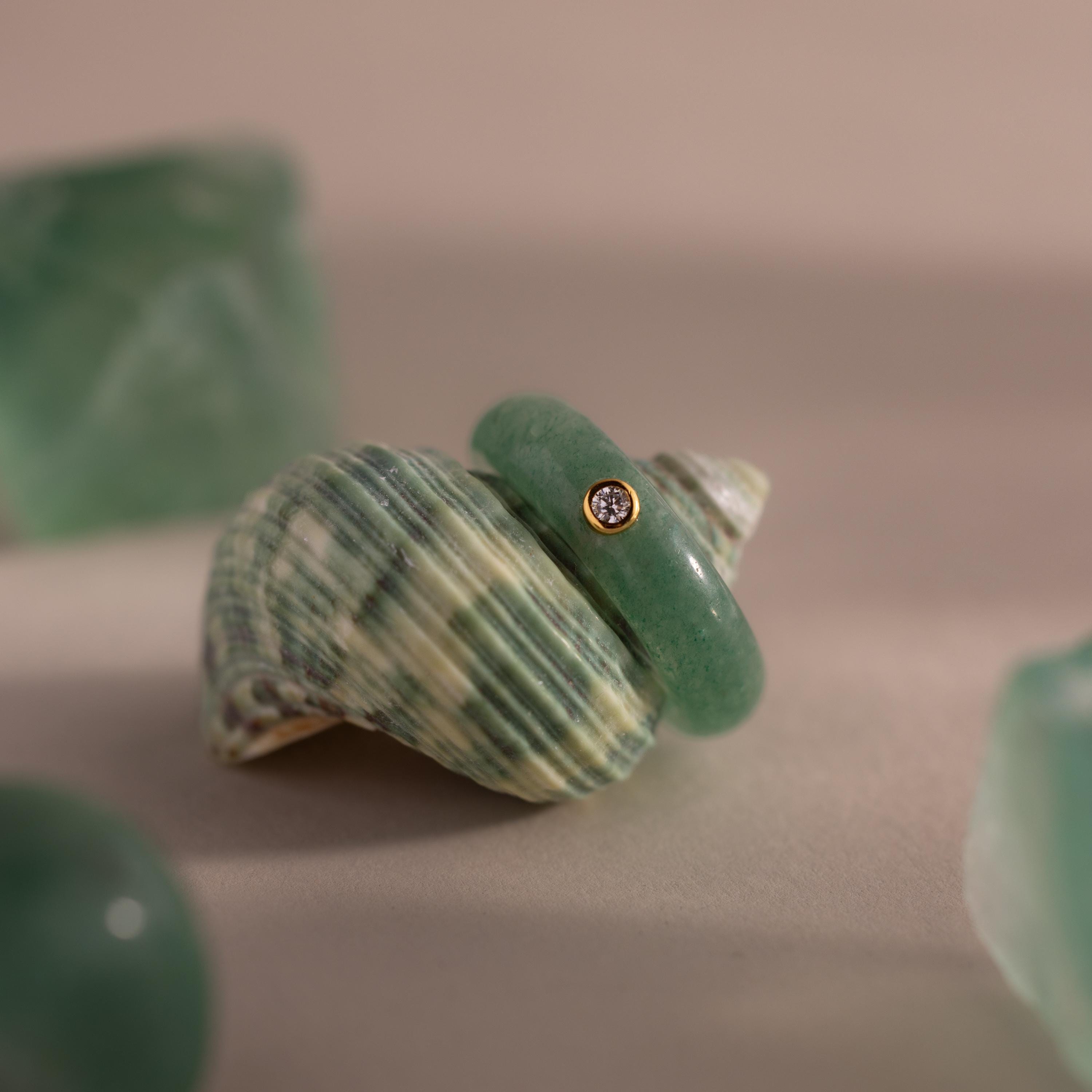 The Jade Diamond Ring features a green jade gemstone and a small diamond set in gold, displayed on a striped seashell with surrounding green stones.