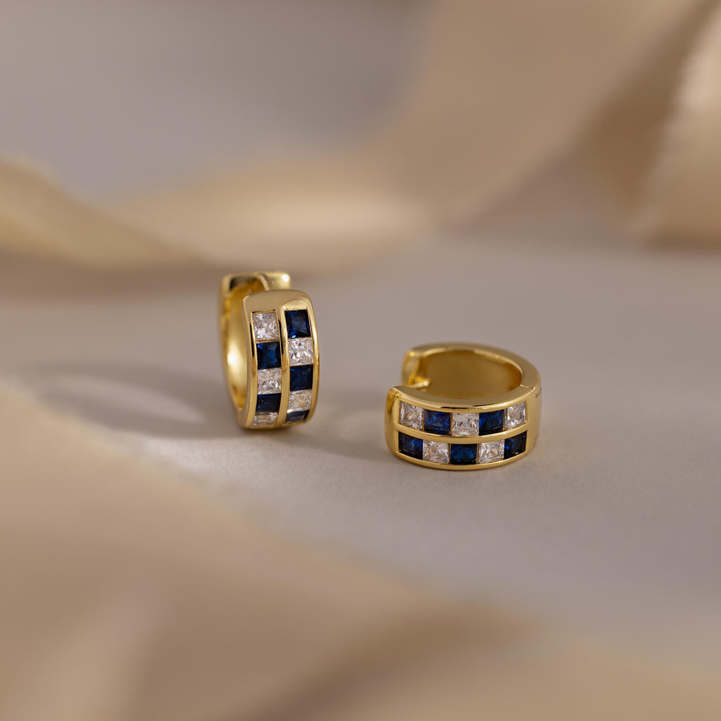 A pair of Royal Blue Checkered Huggies featuring gold geometric design with alternating royal blue and clear square gemstones, displayed on a neutral surface.