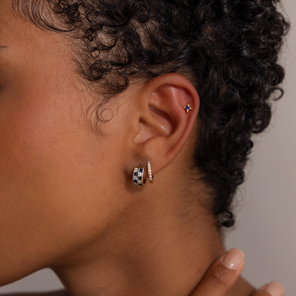 Close-up of a model's ear with three gold earrings, including the Royal Blue Checkered Huggies in 18K Gold and a star-shaped stud.