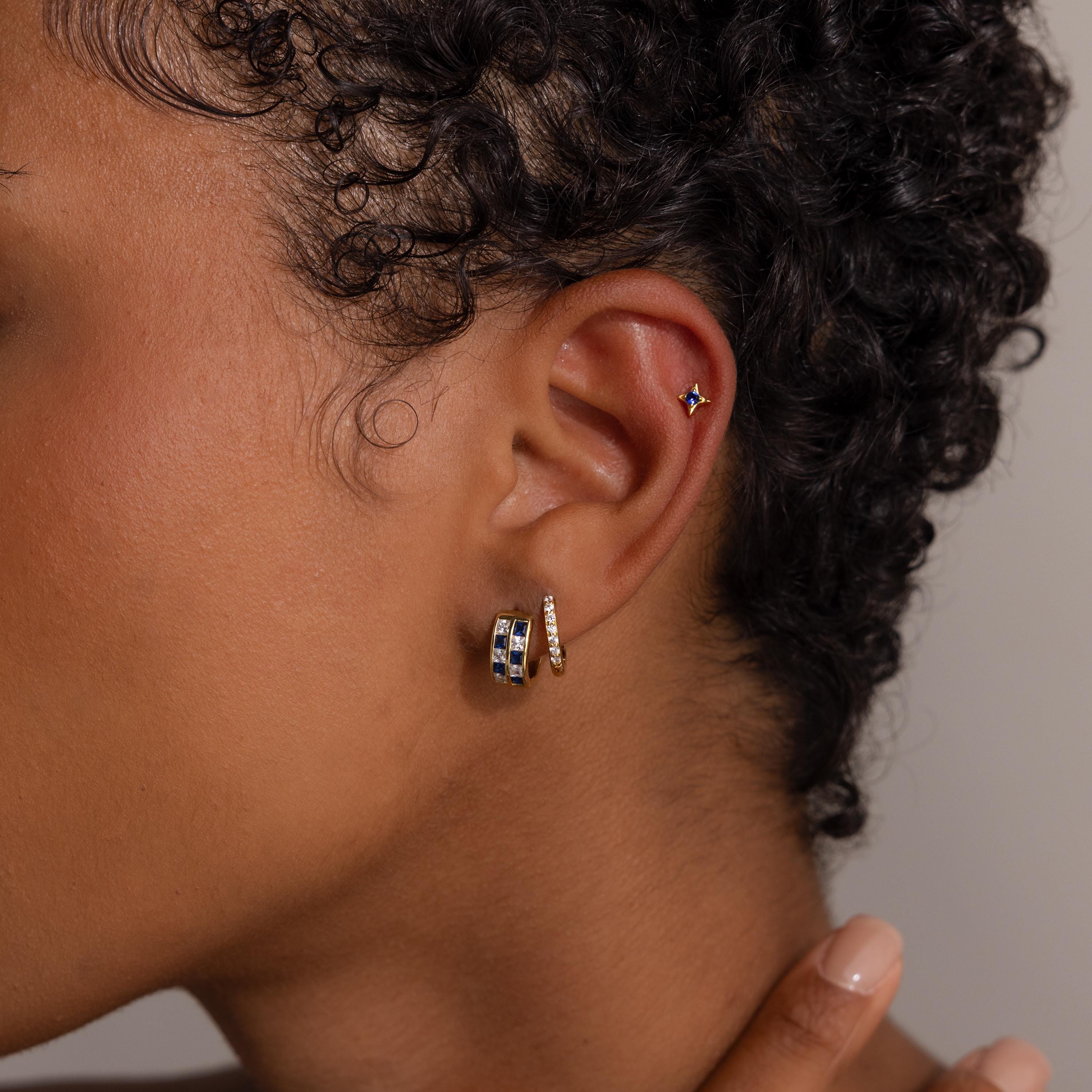 Close-up of a model's ear with three gold earrings, including the Royal Blue Checkered Huggies in 18K Gold and a star-shaped stud.
