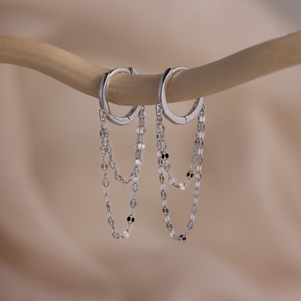 The Duo Mirror Chain Huggies in Sterling Silver—sleek silver hoops with double chain dangles—are displayed on a curved branch, creating an effortlessly stylish, layered earrings look against a soft background.