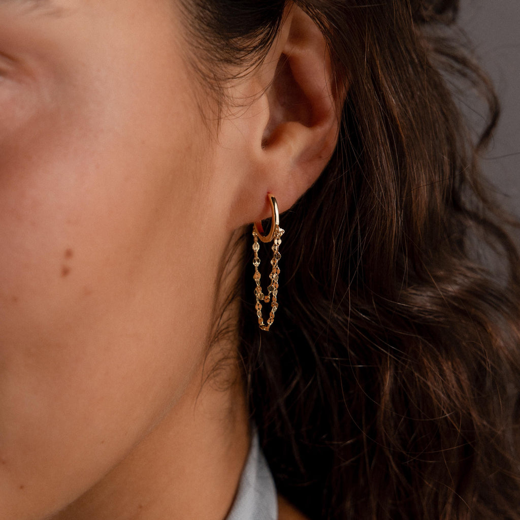 Close-up of a woman's ear wearing Duo Mirror Chain Huggies—modern gold hoops featuring dangling chains and small beads.