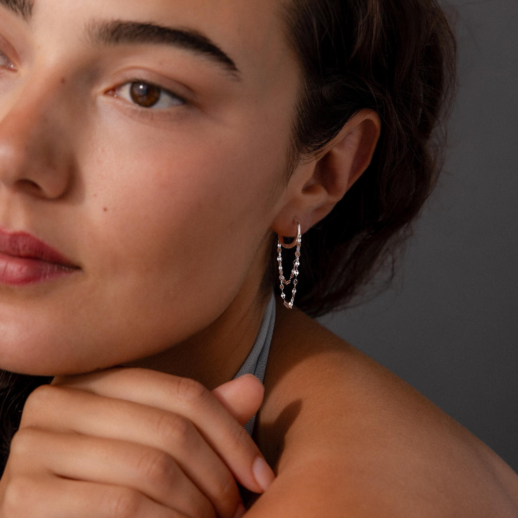 Woman with wavy hair wearing modern jewelry—silver hoop earrings styled as Duo Mirror Chain Huggies—rests her chin on her hand, gazing thoughtfully to the side.