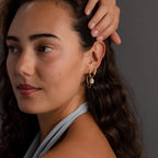A woman with wavy hair touches her head and looks to the side, wearing modern jewelry—a pair of Duo Mirror Chain Huggies styled as layered earrings.