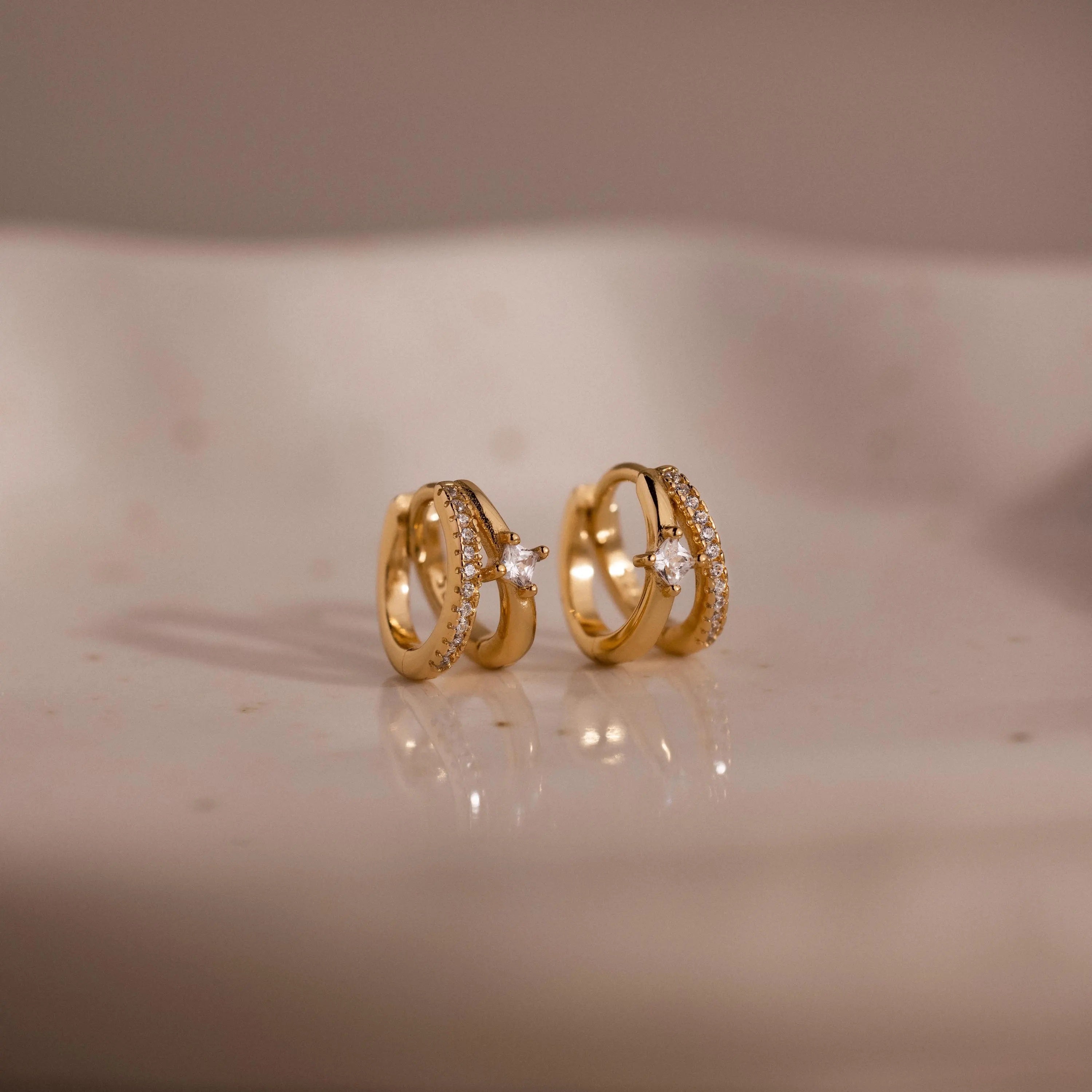 The Pave Diamond Duo Huggies in 18K Gold, featuring elegant gold hoops with small diamonds, are beautifully displayed on a soft, cream-colored surface.