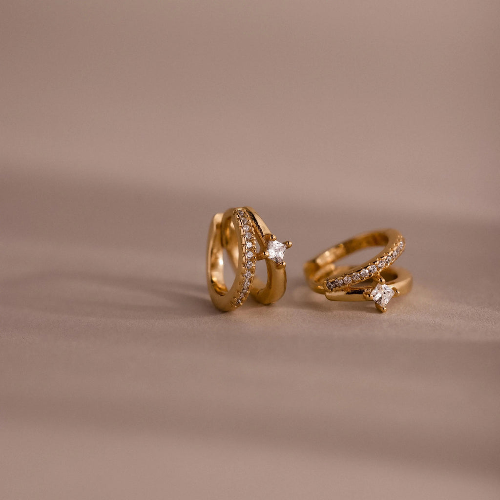 The Pave Diamond Duo Huggies, featuring sparkling diamonds in a gold setting, rest on a soft beige surface with a blurred background.