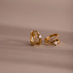 The Pave Diamond Duo Huggies, featuring sparkling diamonds in a gold setting, rest on a soft beige surface with a blurred background.