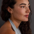 Woman with long curly hair and gold earrings, wearing a light gray halter top and Pave Diamond Duo Huggies, looking to the side.