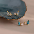 Two pairs of Marquise Birthstone & Pearl Studs, featuring blue and clear stones, are displayed next to a blue ceramic dish.