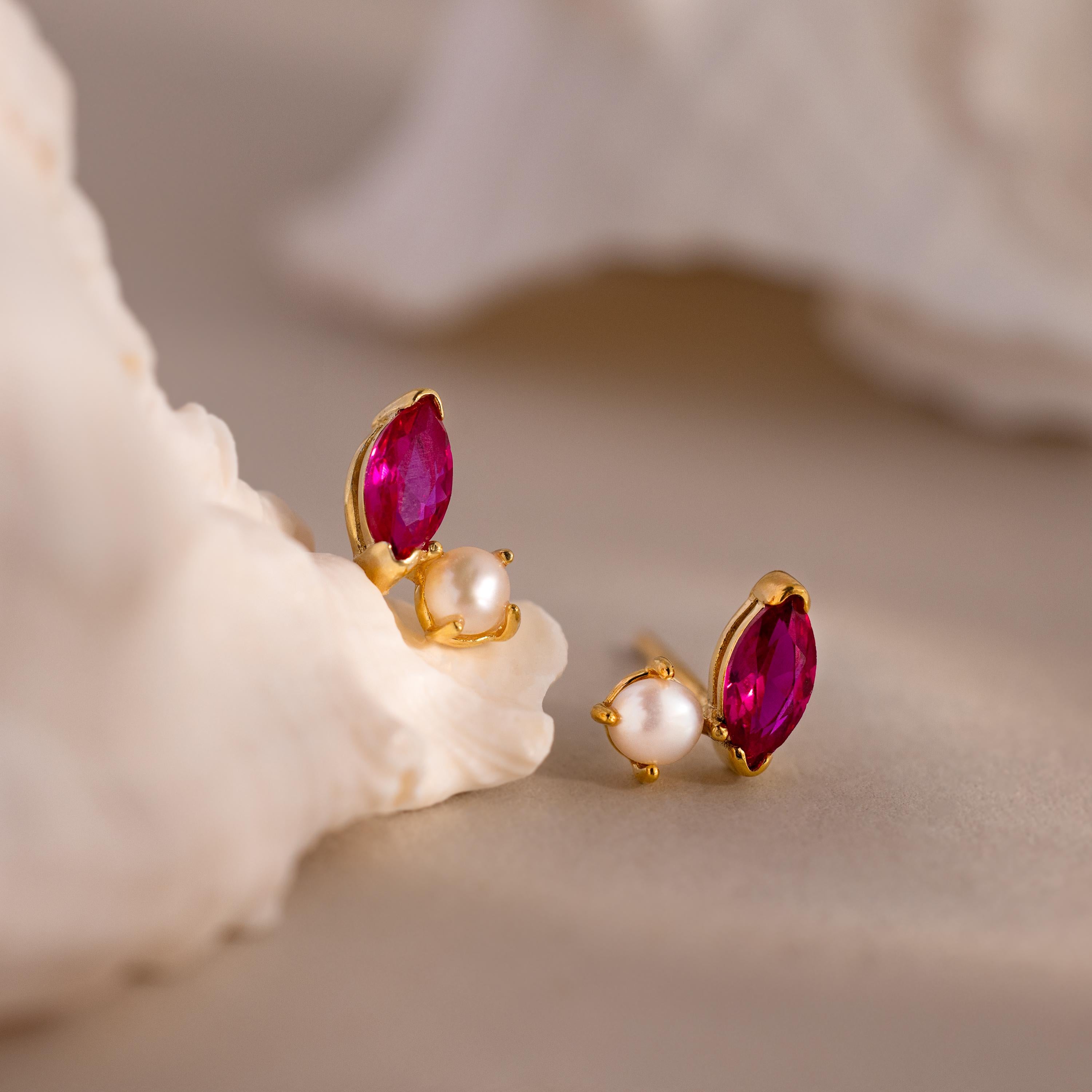 Marquise Ruby & Pearl Studs featuring vibrant red gemstones and white pearls are displayed on a soft, neutral background decorated with shells.