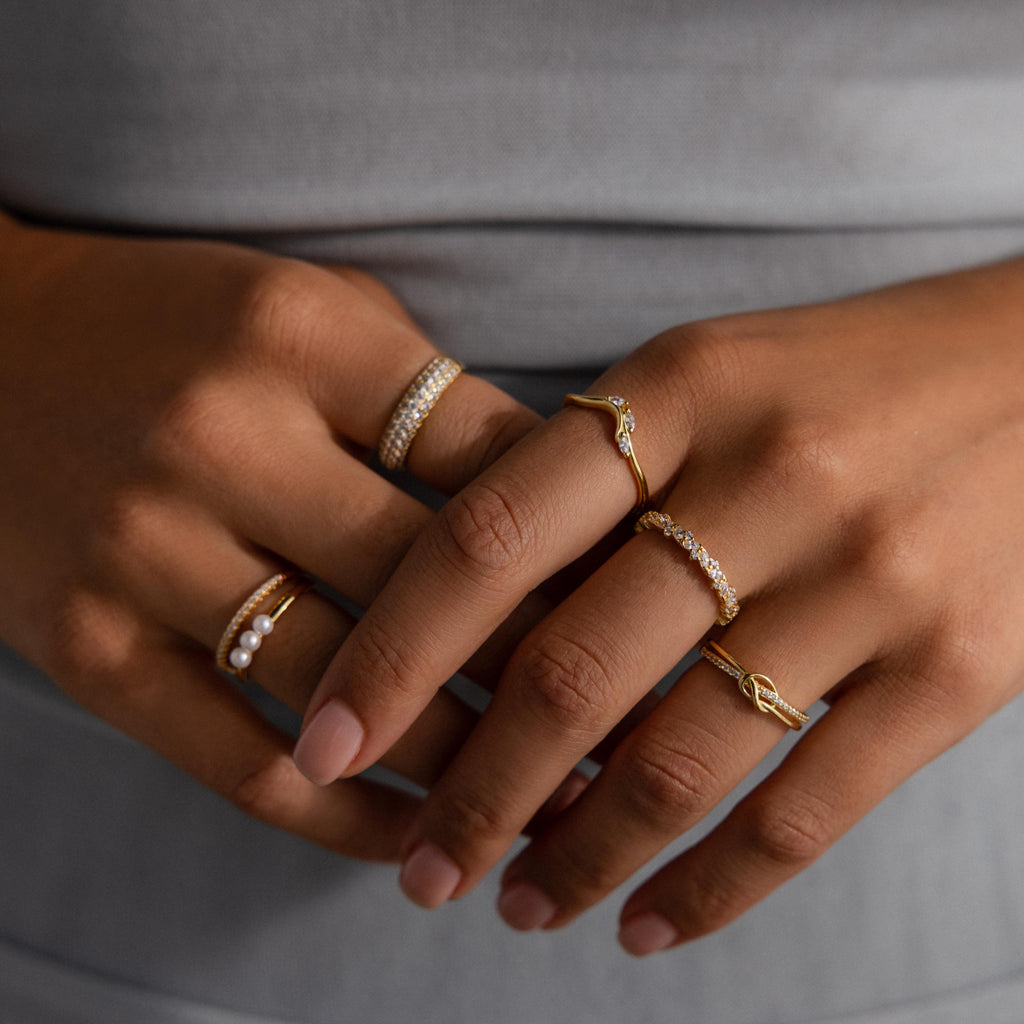 Close-up of hands wearing multiple delicate gold rings, including the Diamond Cluster Eternity Ring, against a gray outfit.