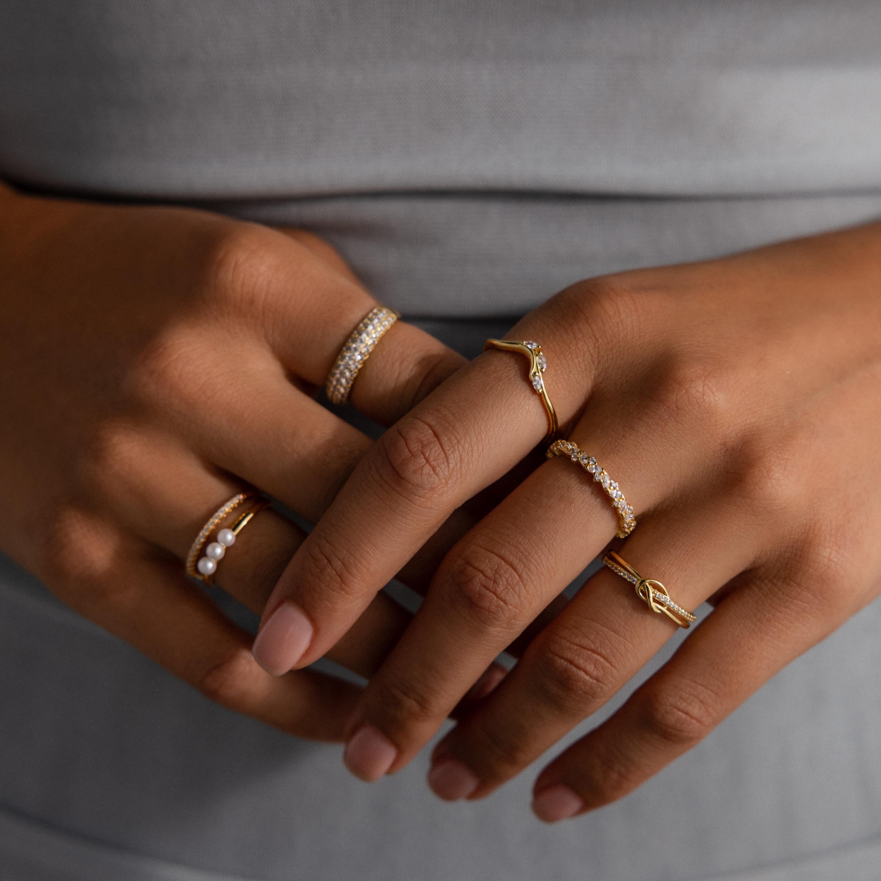 Close-up of hands wearing multiple delicate gold rings, including the Diamond Cluster Eternity Ring, against a gray outfit.