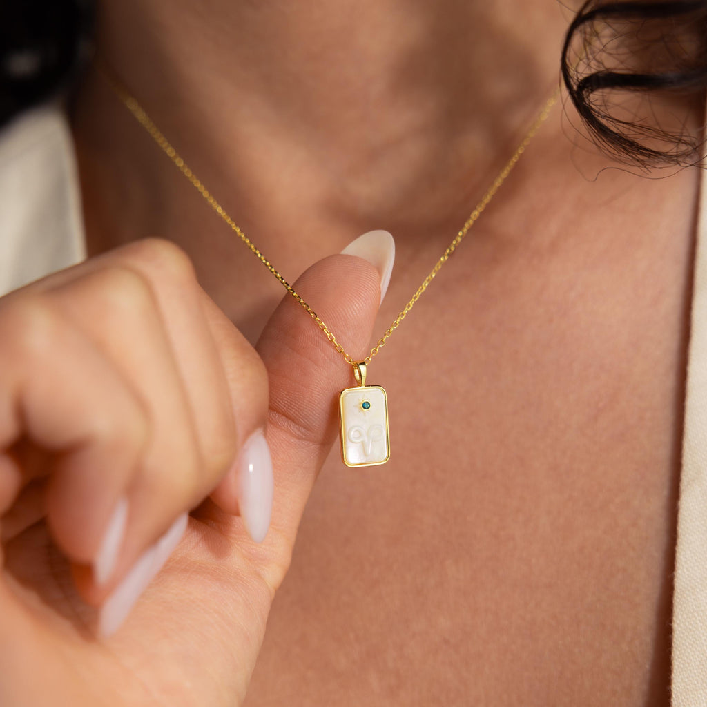 A hand holds the Pearl Zodiac Birthstone Necklace in 18K Gold, featuring a rectangular pendant with intricate designs, worn around the neck.