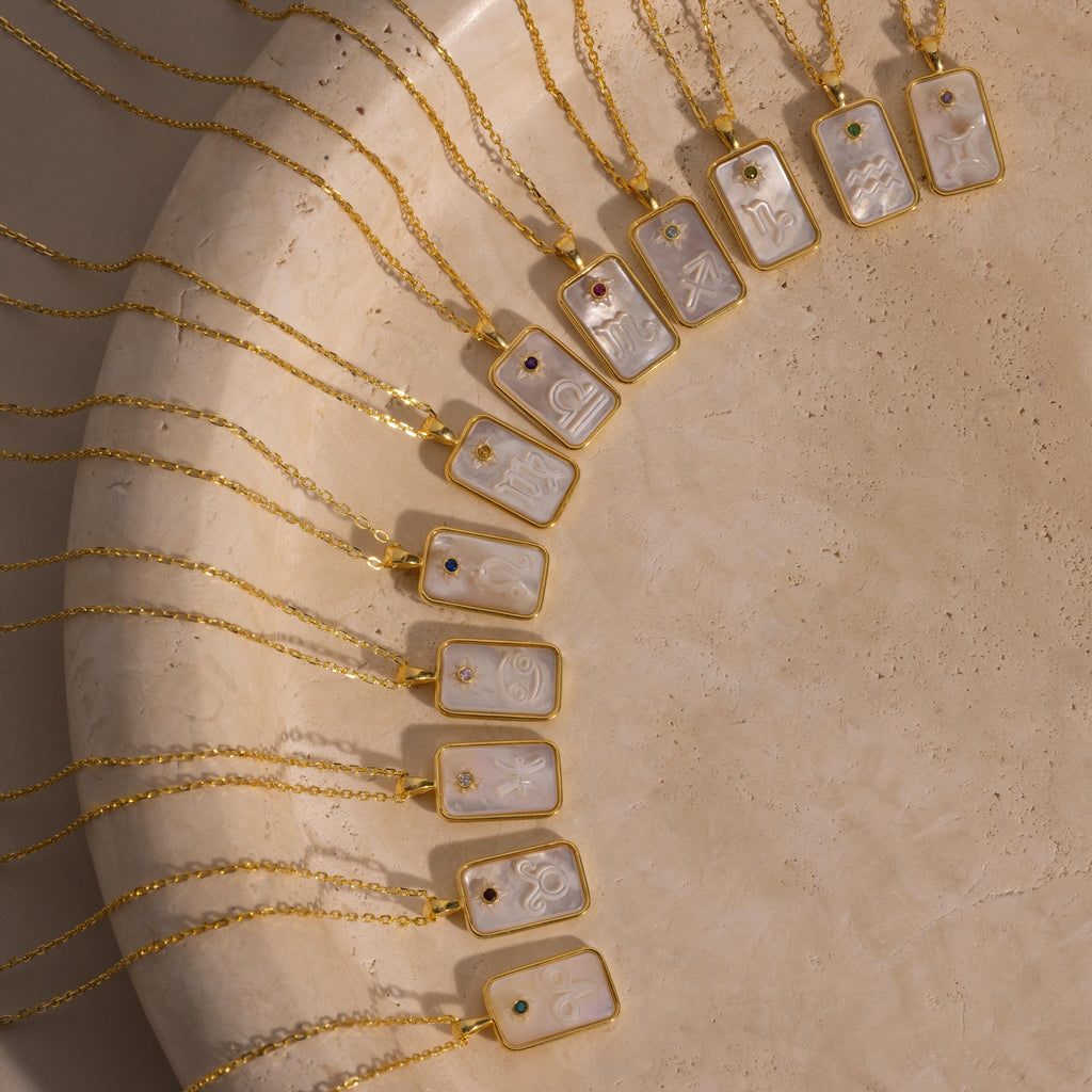 The Pearl Zodiac Birthstone Necklace in Sterling Silver is displayed with other rectangular pendant zodiac jewelry, arranged in a half-circle on a textured beige surface.