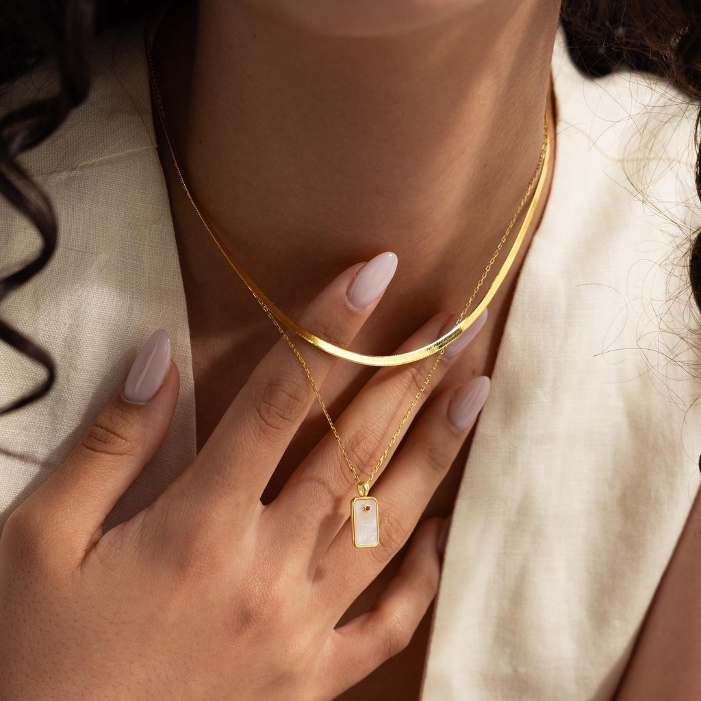 A woman in a cream top touches her chest, showing off layered gold necklaces, including the Pearl Zodiac Birthstone Necklace in Rose Gold, with manicured nails.