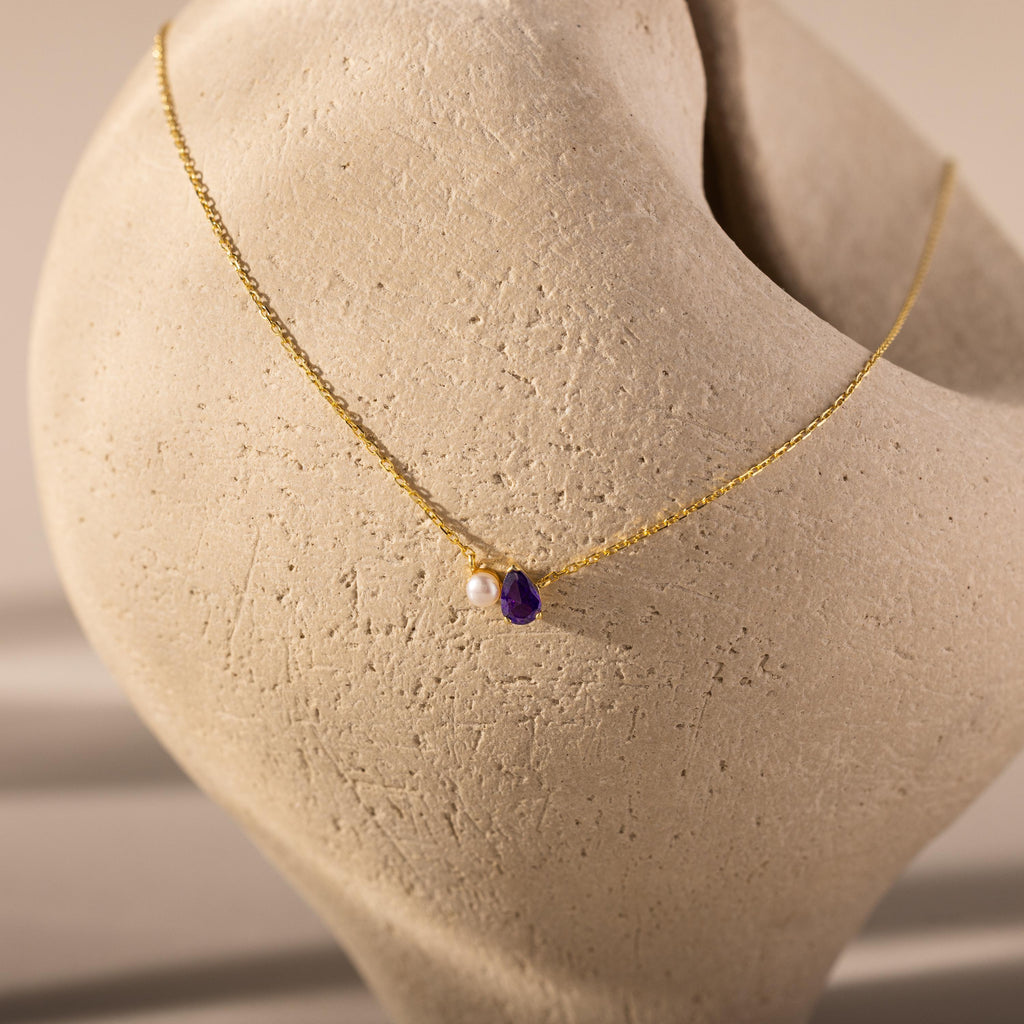 The Teardrop Birthstone & Pearl Necklace, featuring a pearl and birthstone pendant, is elegantly draped over a textured beige sculpture—a perfect milestone gift.