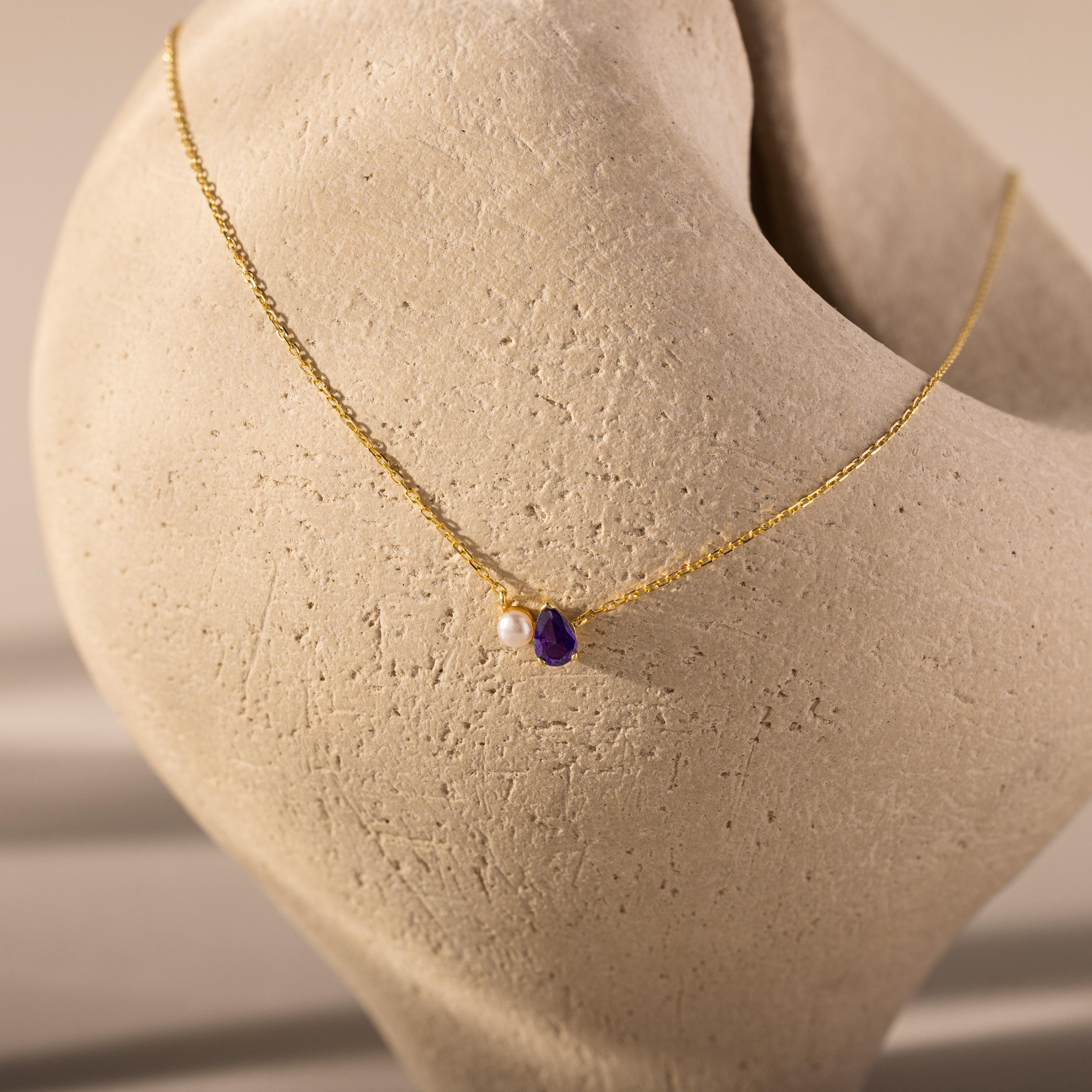 The Teardrop Birthstone & Pearl Necklace, featuring a pearl and birthstone pendant, is elegantly draped over a textured beige sculpture—a perfect milestone gift.