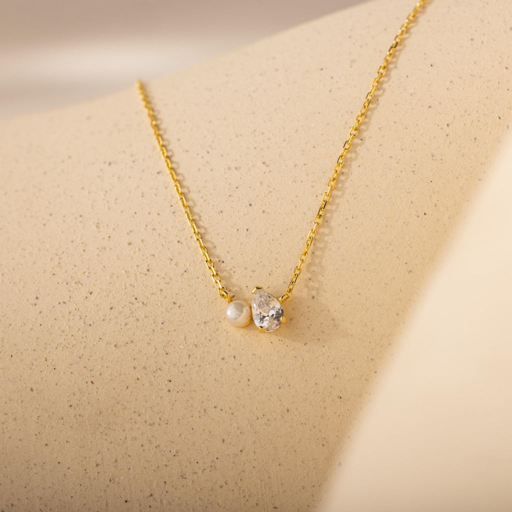 The Teardrop Diamond & Pearl Necklace features a pearl and crystal pendant on a gold chain, shown on a beige textured surface—an elegant gift for any occasion.