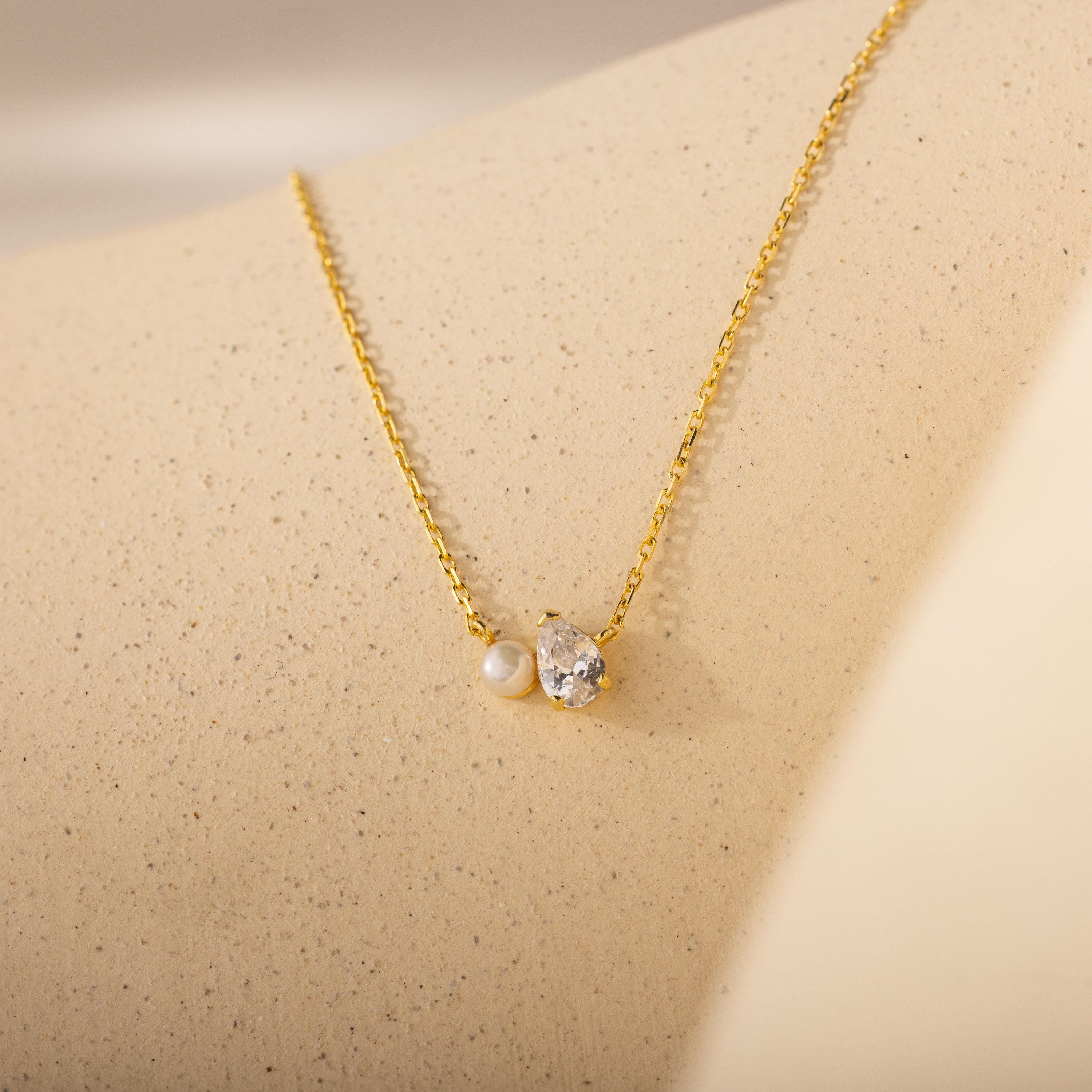 The Teardrop Diamond & Pearl Necklace features a pearl and crystal pendant on a gold chain, shown on a beige textured surface—an elegant gift for any occasion.