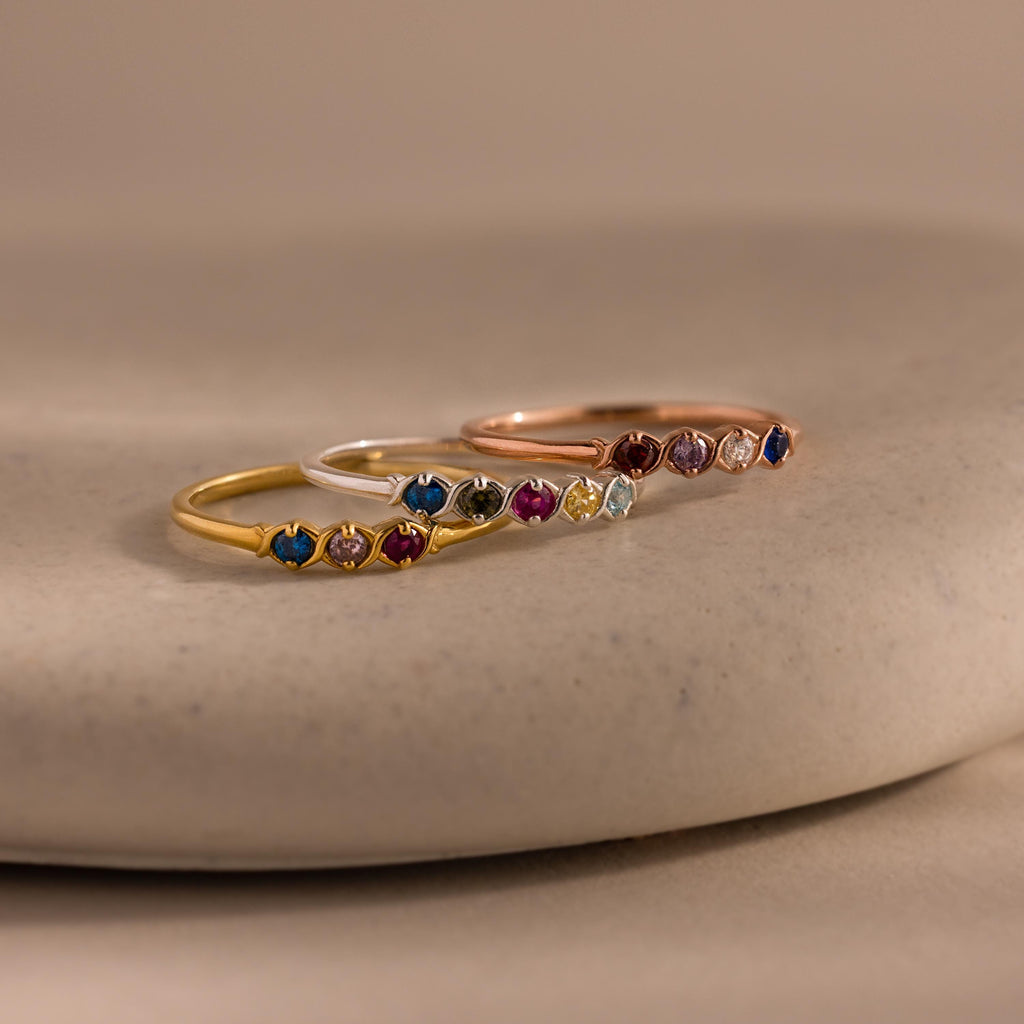 The Woven Multiple Birthstone Ring set features three delicate, vintage-inspired rings in gold, silver, and rose gold with colorful gemstones, beautifully displayed on a smooth beige surface.
