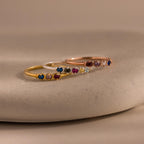 The Woven Multiple Birthstone Ring set features three delicate, vintage-inspired rings in gold, silver, and rose gold with colorful gemstones, beautifully displayed on a smooth beige surface.