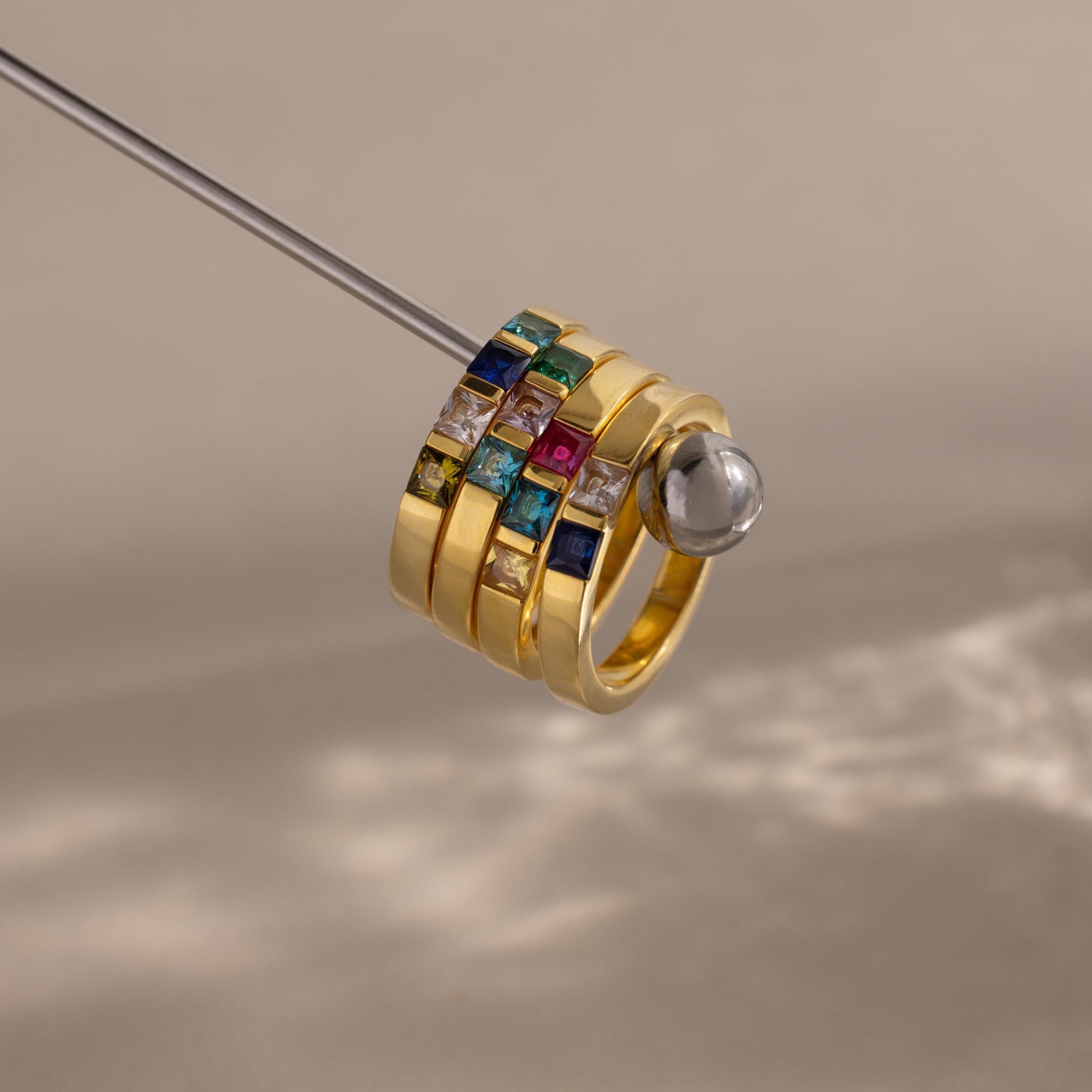 The Multiple Birthstone Band in 18K Gold features four stacked gold rings set with colorful square gemstones, creating a stunning personalized jewelry piece against a neutral background.