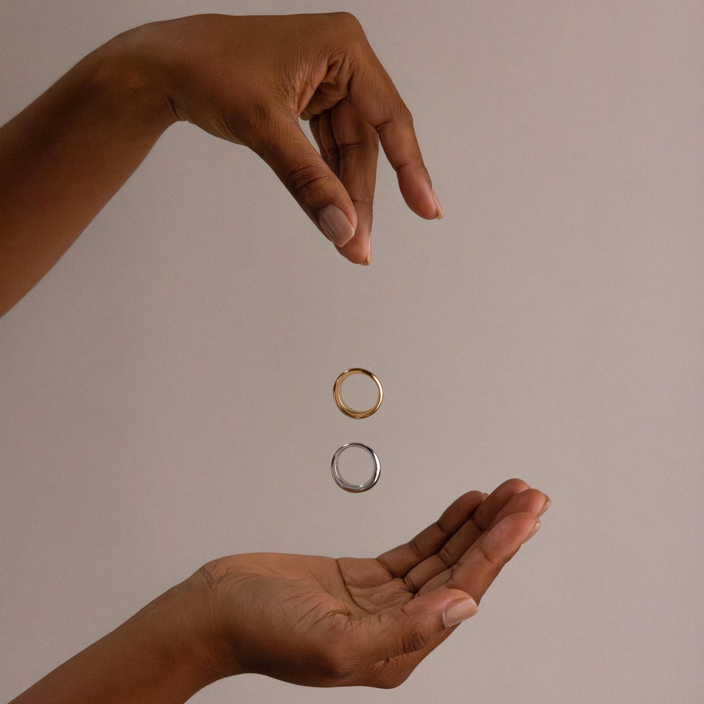 Two hands frame a floating gold ring above a silver ring, highlighting the Mixed Metal Dome Ring Set—a versatile and modern jewelry collection—against a plain beige background.
