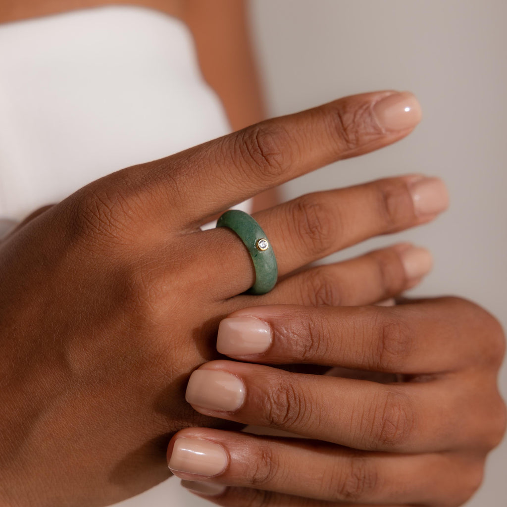 A hand with nude nails displays the Jade Diamond Ring—a green jade stone with a small diamond set in a gold bezel—on the ring finger.