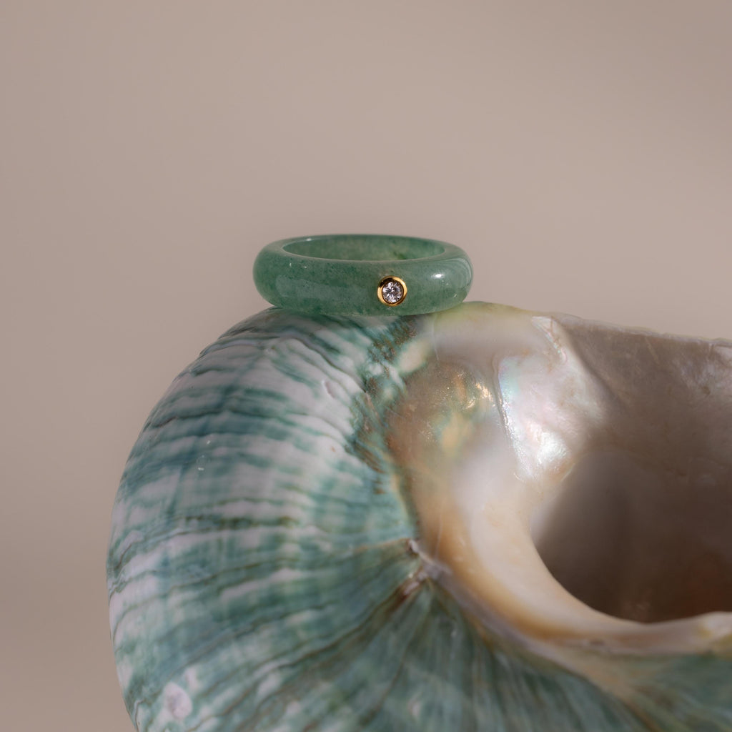 The Jade Diamond Ring features a small diamond in a gold bezel, displayed on a green and white seashell against a neutral background.