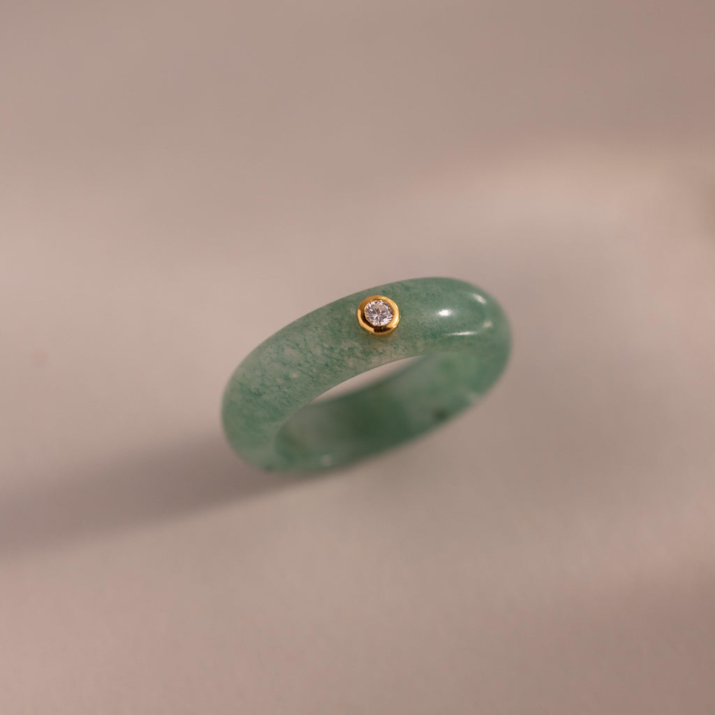 The Jade Diamond Ring features green jade set in a gold bezel with a single clear diamond, displayed on a neutral background.