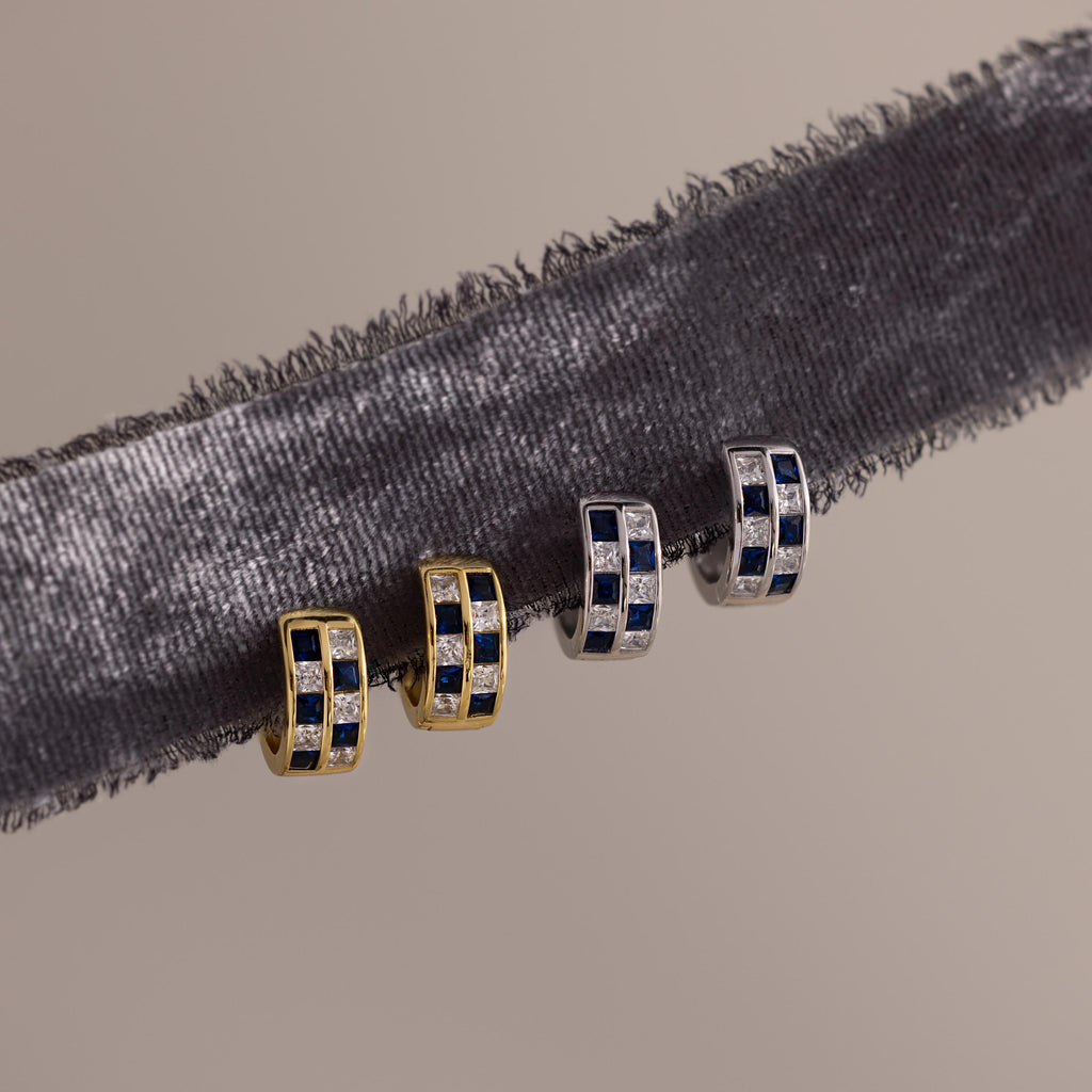 Two pairs of earrings, featuring gold geometric styles and Royal Blue Checkered Huggies with silver, blue, and clear stones, are displayed on a velvet ribbon.