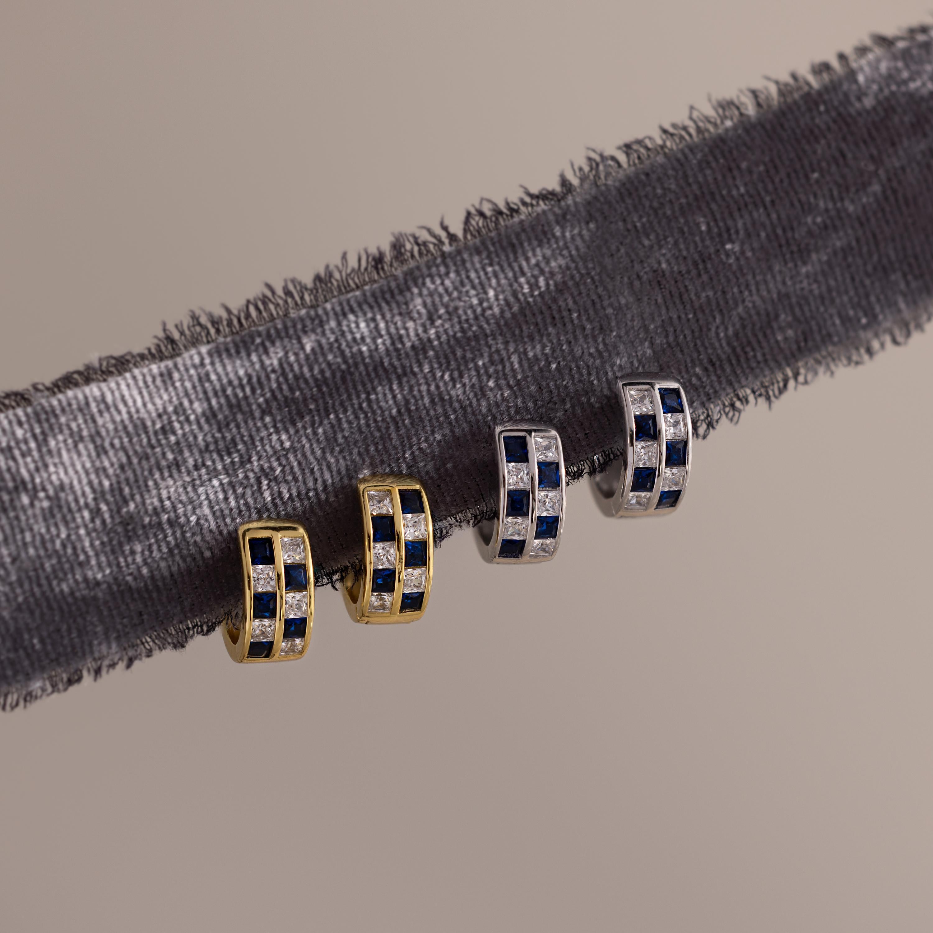 Two pairs of earrings, featuring gold geometric styles and Royal Blue Checkered Huggies with silver, blue, and clear stones, are displayed on a velvet ribbon.