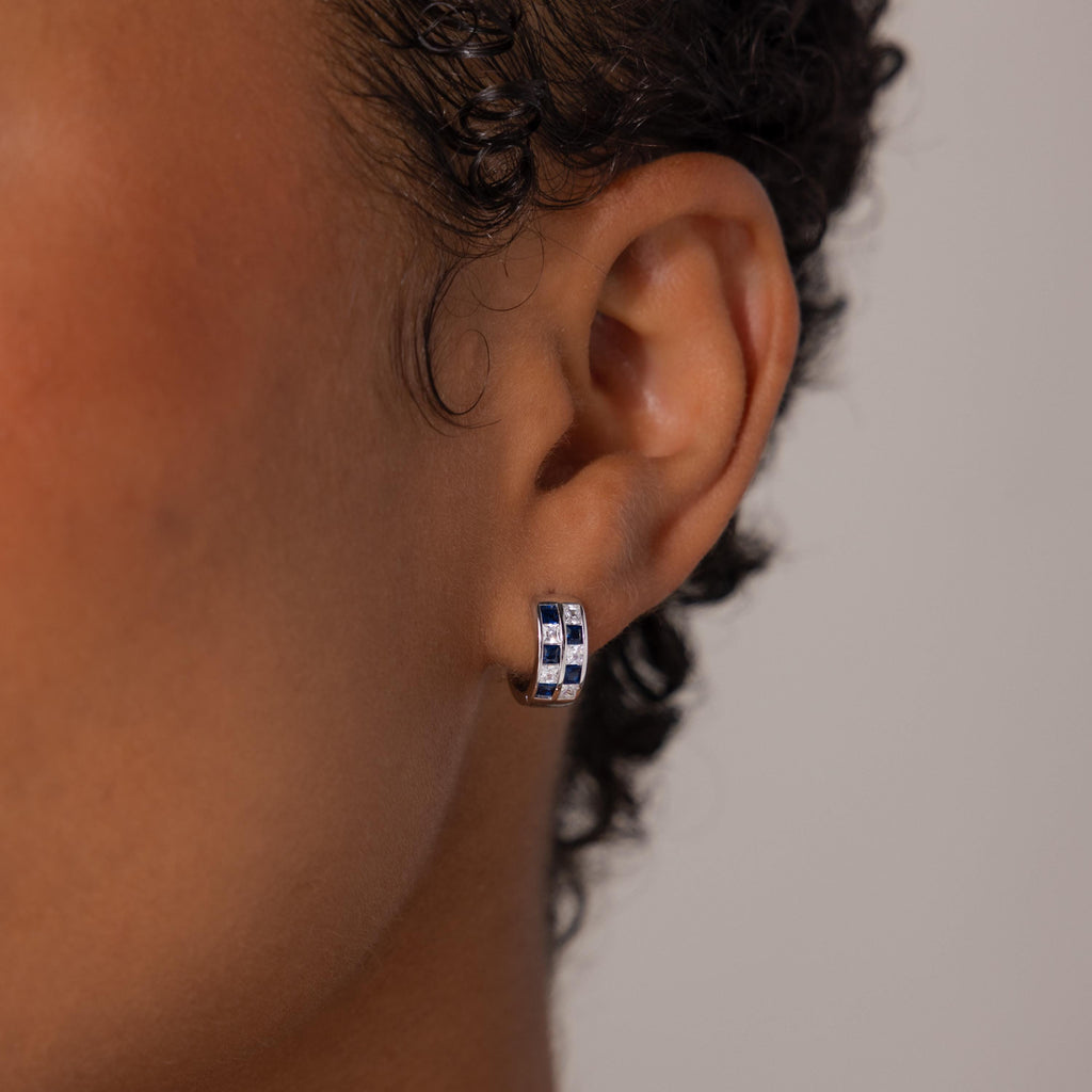 Close-up of a person's ear wearing Royal Blue Checkered Huggies in Sterling Silver, featuring blue and clear stones.