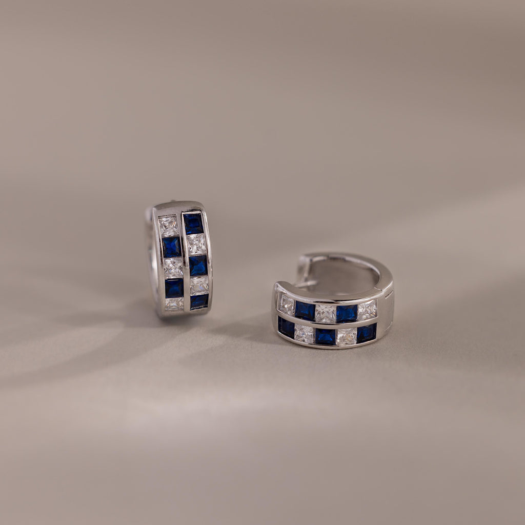 The Royal Blue Checkered Huggies in Sterling Silver feature alternating blue and clear gemstones in a chic, geometric pattern, shown on a neutral background for a stylish and modern touch.