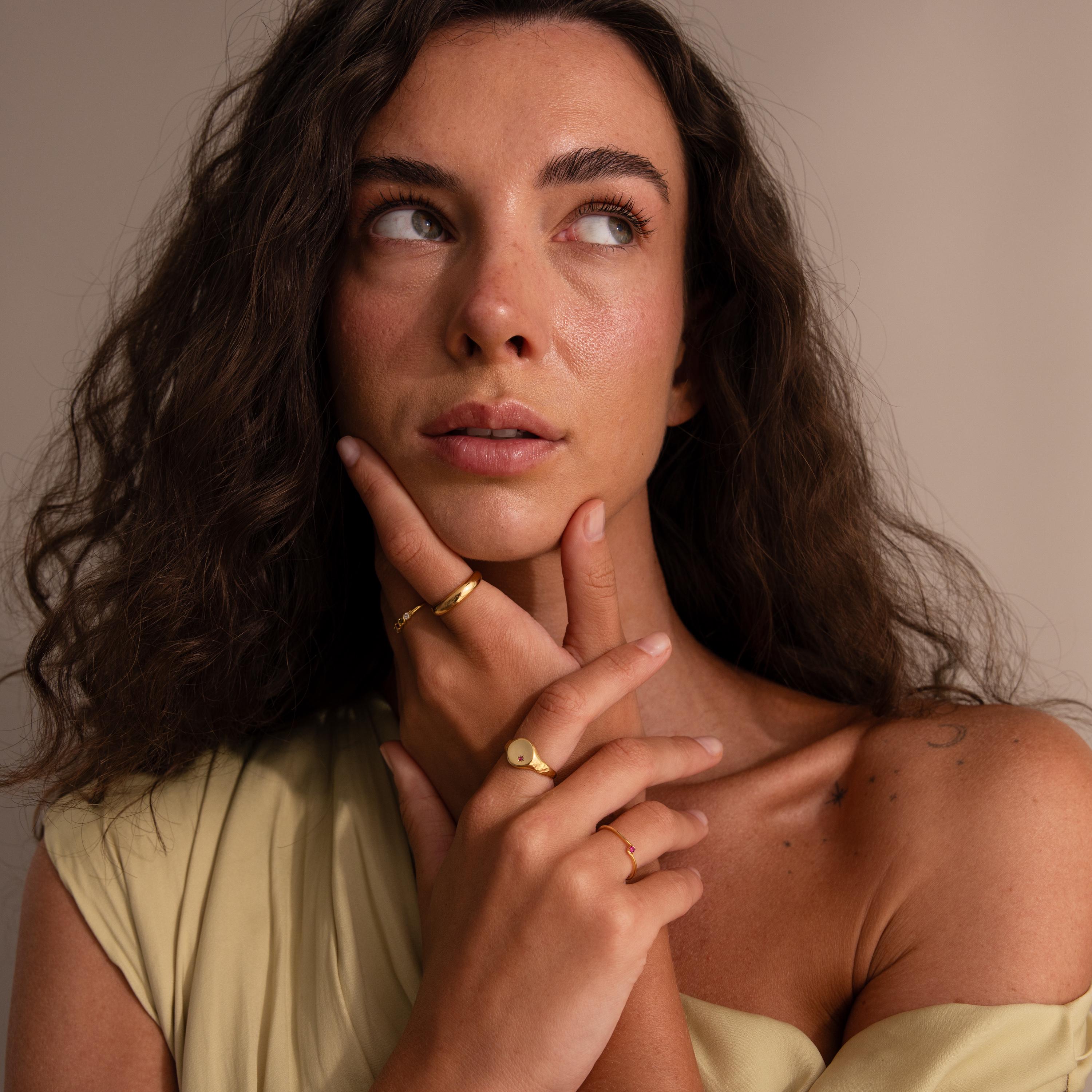 A woman with wavy brown hair wears the Woven Multiple Birthstone Ring and gold bands, posing thoughtfully with her hand on her chin against a neutral background.