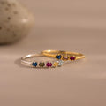 Two delicate rings—a gold vintage-style band and the Woven Multiple Birthstone Ring in silver, adorned with colorful gemstones—are displayed on a neutral background.