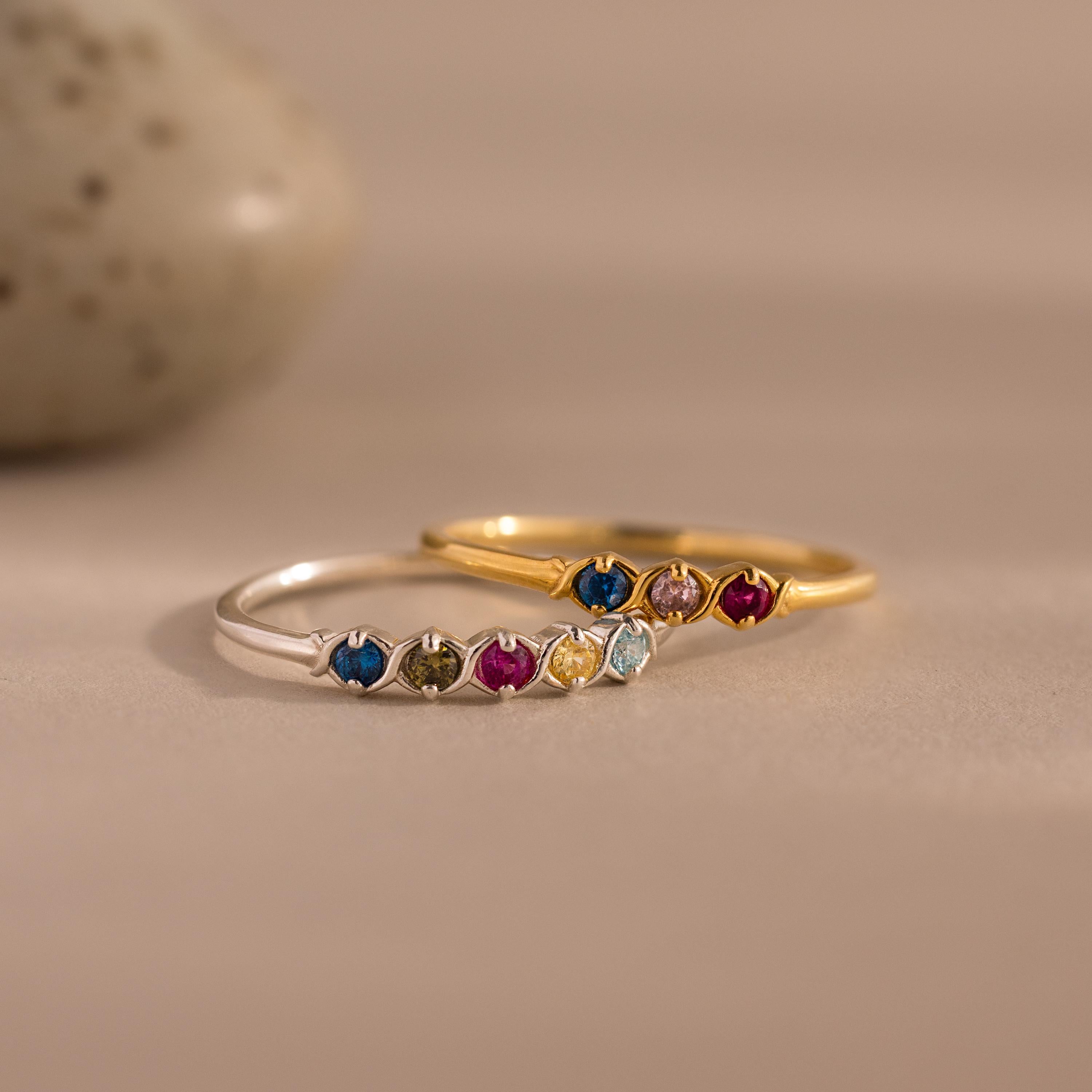 Two delicate rings—a gold vintage-style band and the Woven Multiple Birthstone Ring in silver, adorned with colorful gemstones—are displayed on a neutral background.