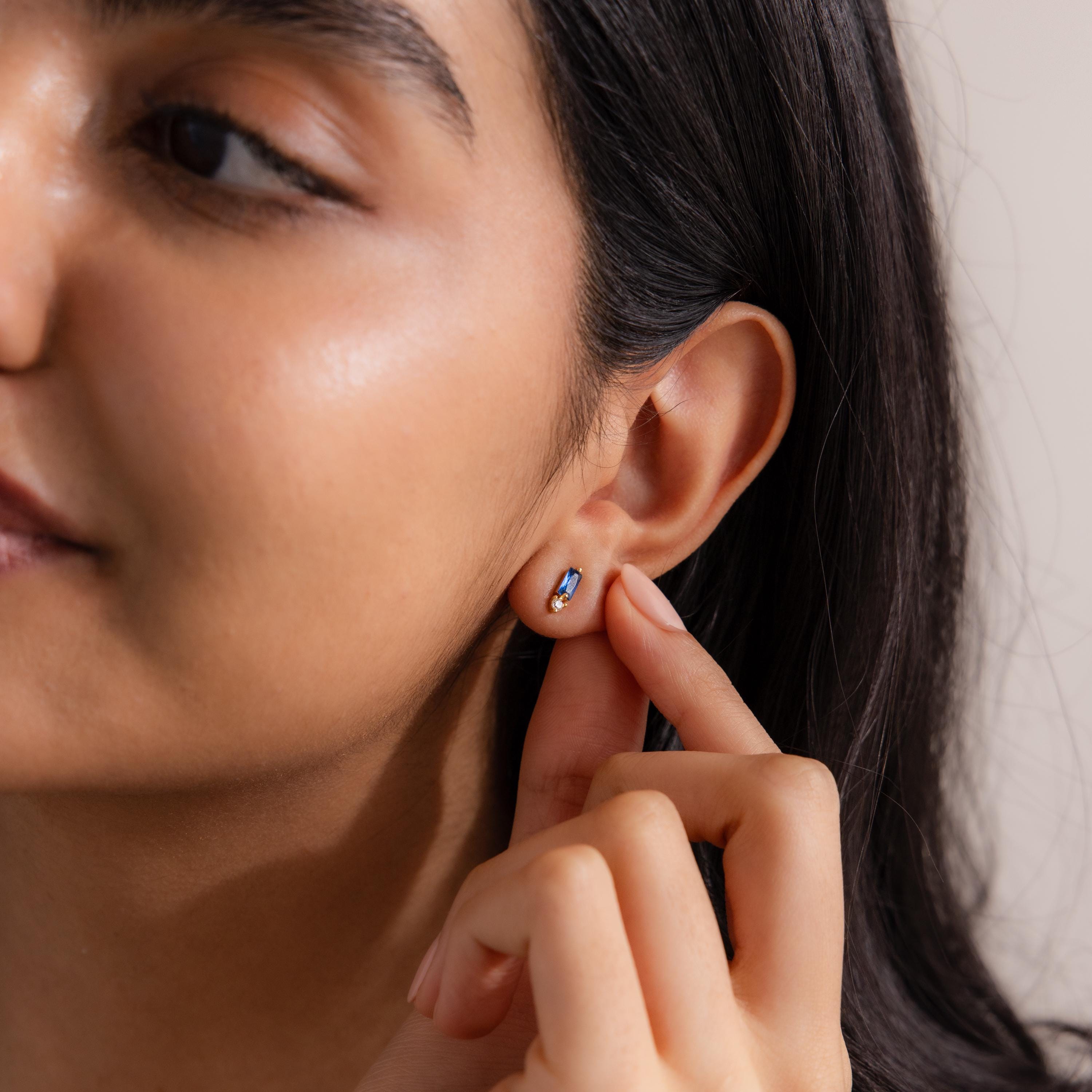 A woman touches a Baguette Blue Zircon Drop Stud on her ear, elegantly showcasing these modern birthstone-style gemstone earrings.