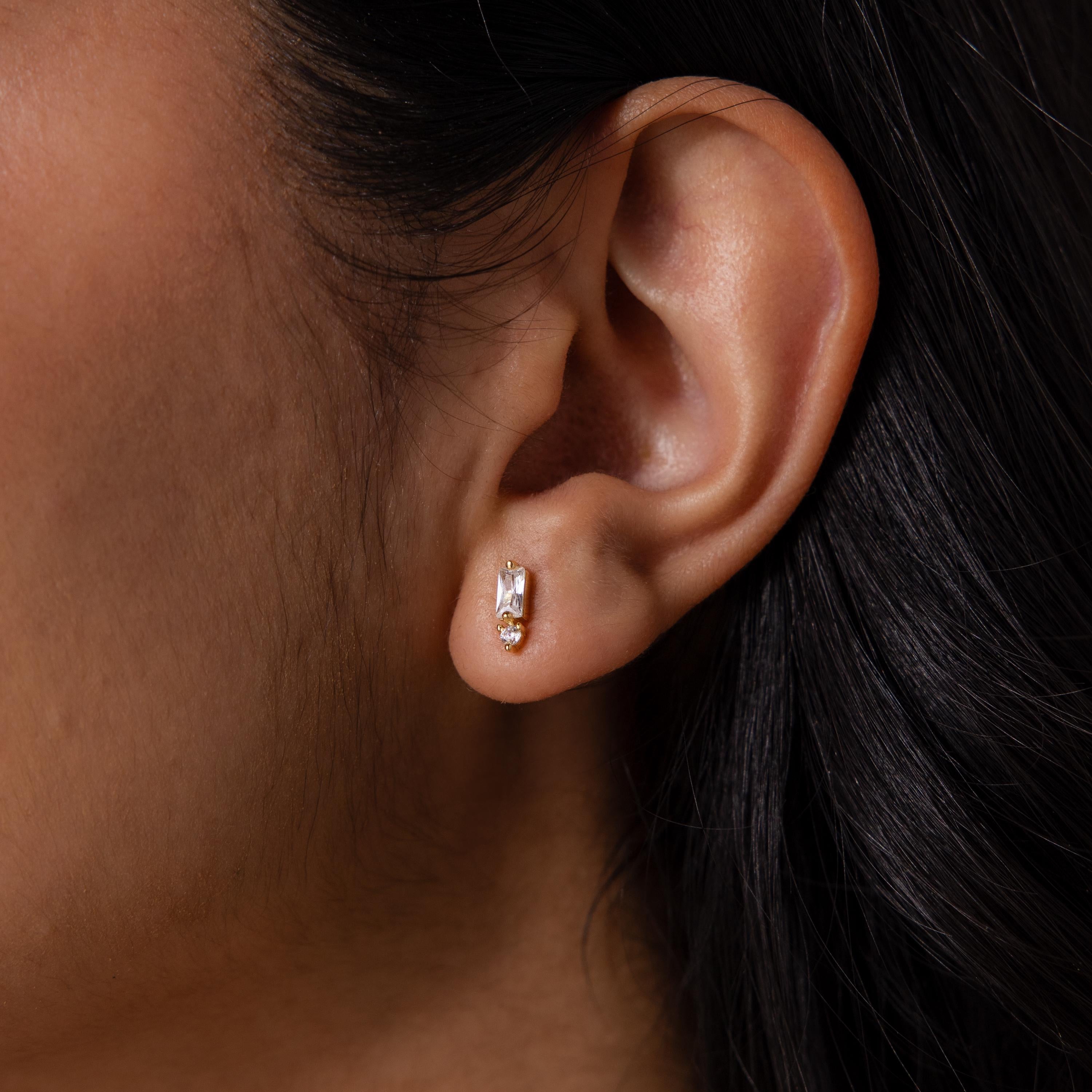 Close-up of a woman's ear wearing Baguette Birthstone Drop Studs, featuring modern rectangular baguette settings with a clear stone and a small round gem.