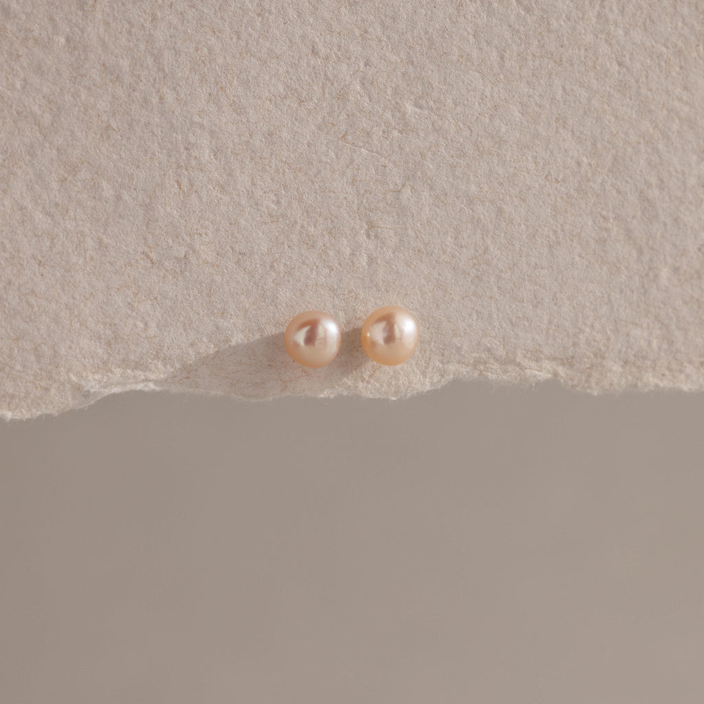 The Pink Pearl Studs, featuring two round blush-toned freshwater pearls, are displayed on a textured beige surface with soft lighting.