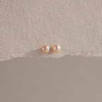 The Pink Pearl Studs, featuring two round blush-toned freshwater pearls, are displayed on a textured beige surface with soft lighting.