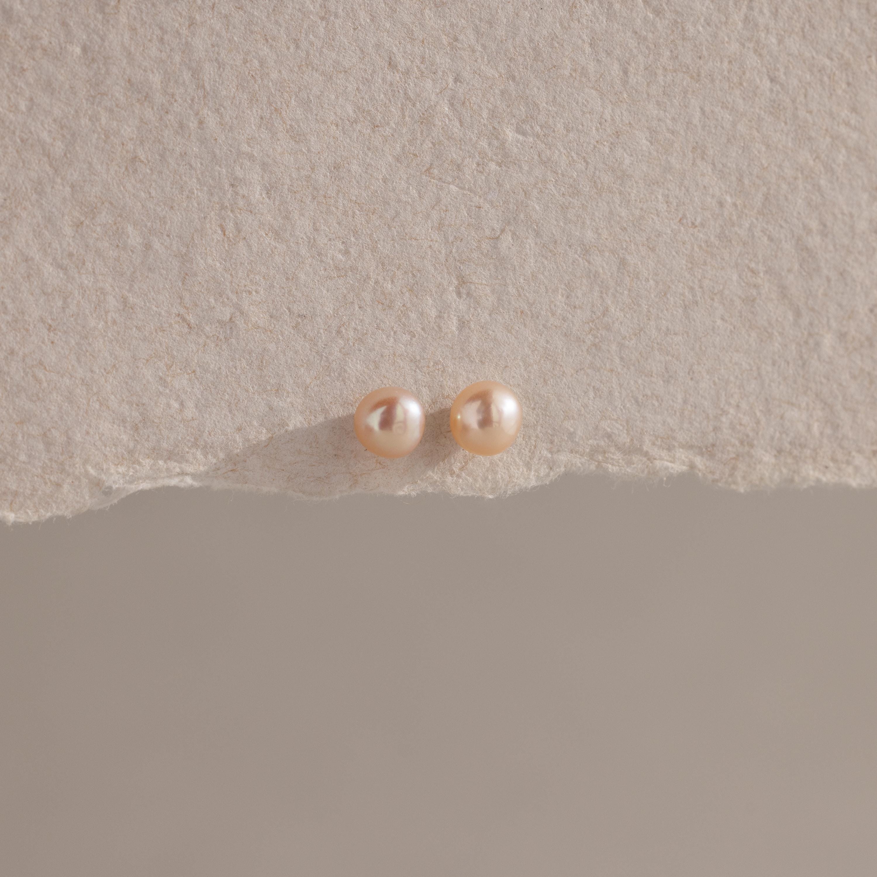 The Pink Pearl Studs, featuring two round blush-toned freshwater pearls, are displayed on a textured beige surface with soft lighting.