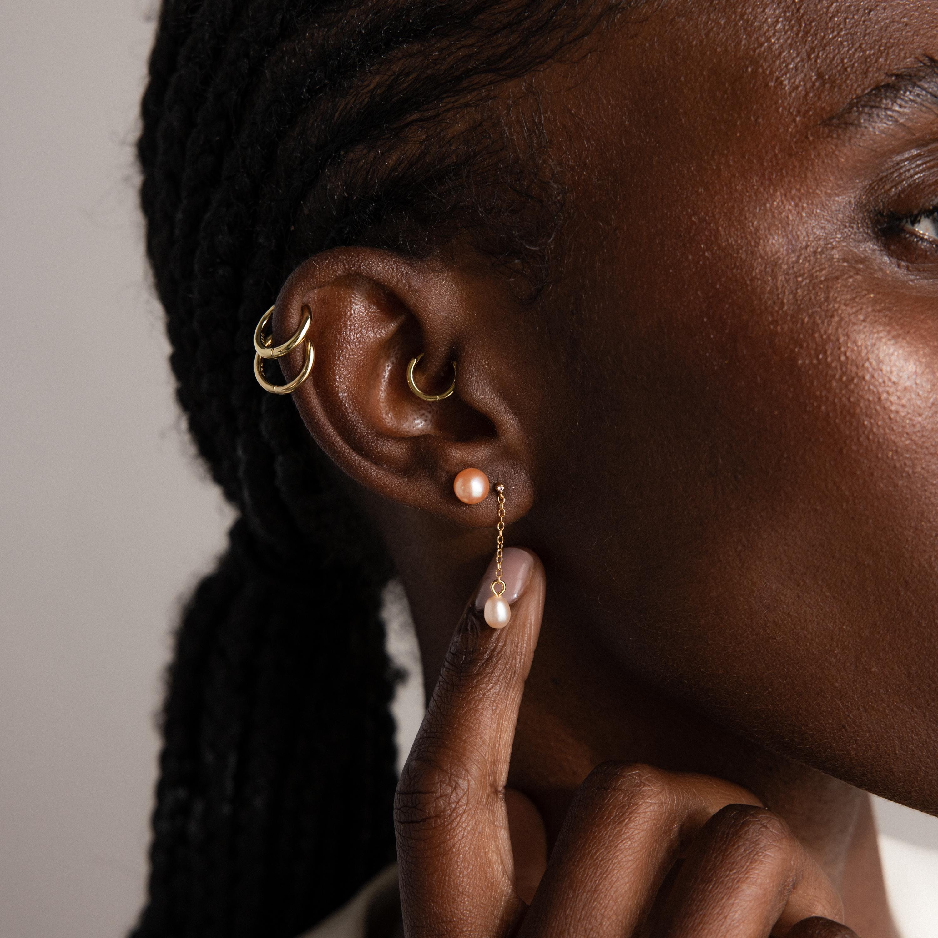 Close-up of a person’s ear wearing Pink Pearl Studs—blush-tone freshwater pearl earrings—alongside gold hoops, with a hand gently touching the jewelry.